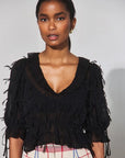 A model wearing a black sheer ruffled blouse with tiered puff sleeves and a deep V-neck, paired with a pink, white, and blue plaid skirt, against a plain light gray background.
