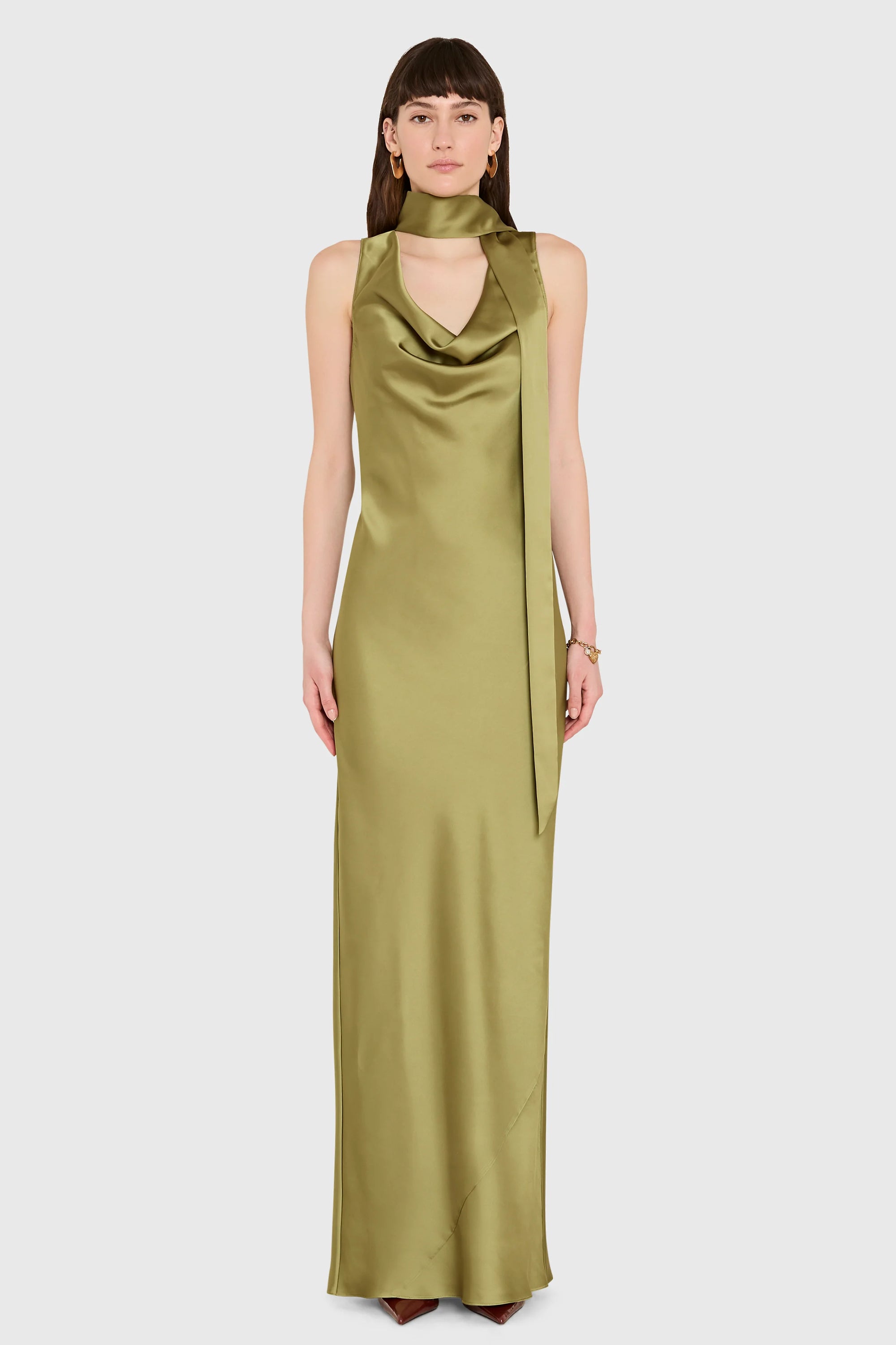 A model wearing a sleeveless, floor-length olive green satin gown with a draped cowl neckline and a matching tie at the waist, set against a solid light gray background.