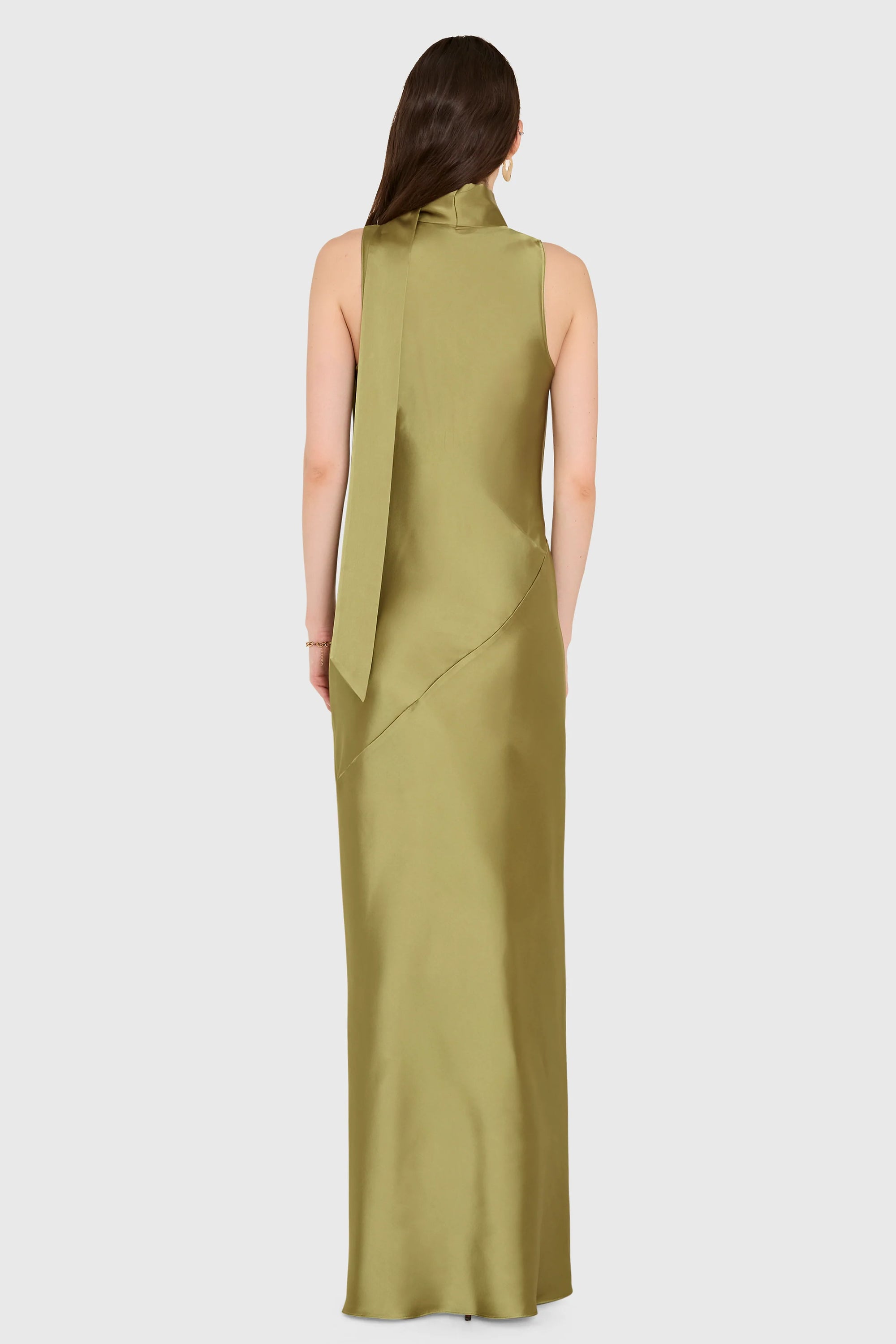 A woman viewed from the back wearing a sleeveless, high-neck, floor-length dress in a muted olive green satin-like fabric against a light gray studio background.