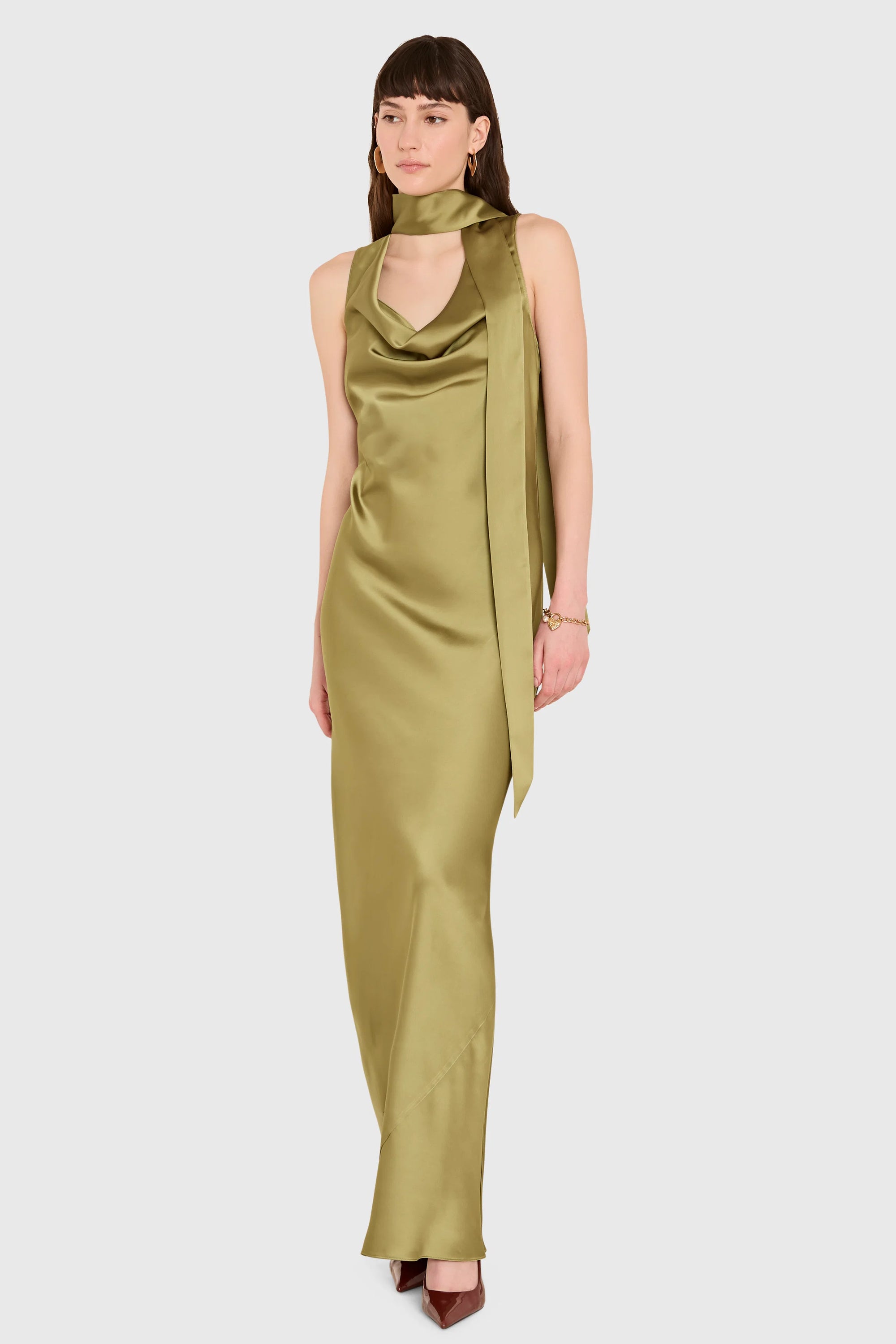 A woman models a sleeveless olive green satin dress with a draped cowl neckline and a long side drape, set against a solid light gray background.