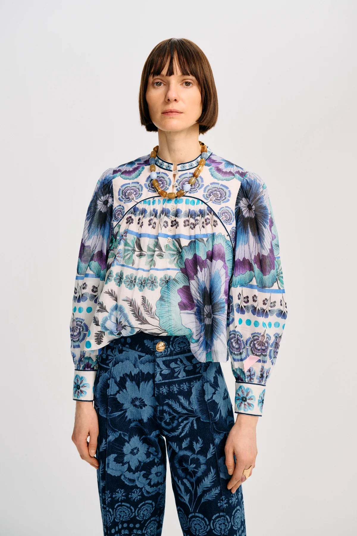 A model wearing a long-sleeve patterned blouse with blue, purple, and white floral motifs, paired with dark blue patterned trousers, against a light gray studio background.