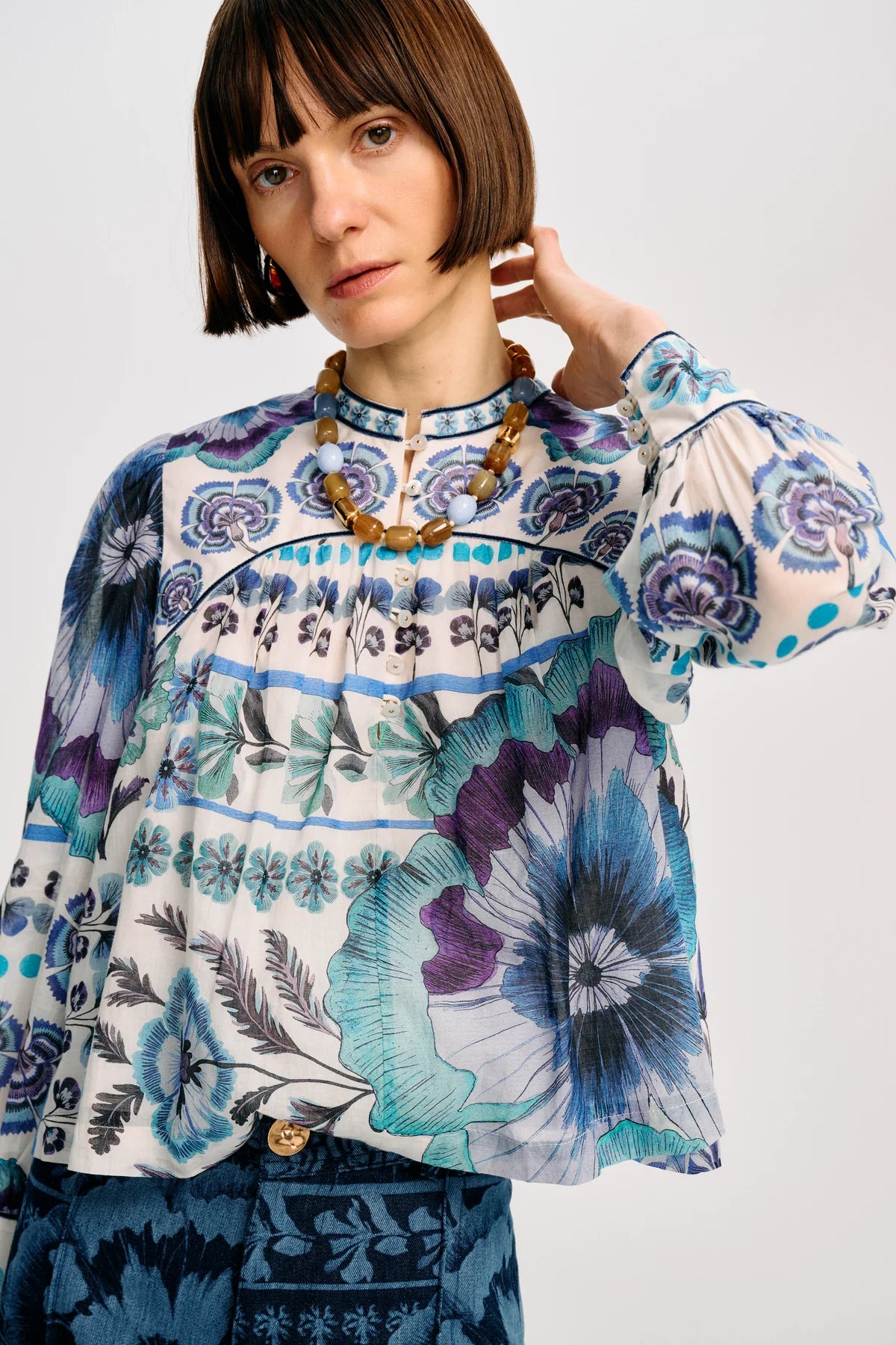 A woman wearing a long-sleeve blouse with a blue, purple, and teal floral print on a white base, paired with a chunky gold necklace, against a light gray studio background.