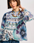 A woman wearing a long-sleeve blouse with a blue, purple, and teal floral print on a white base, paired with a chunky gold necklace, against a light gray studio background.