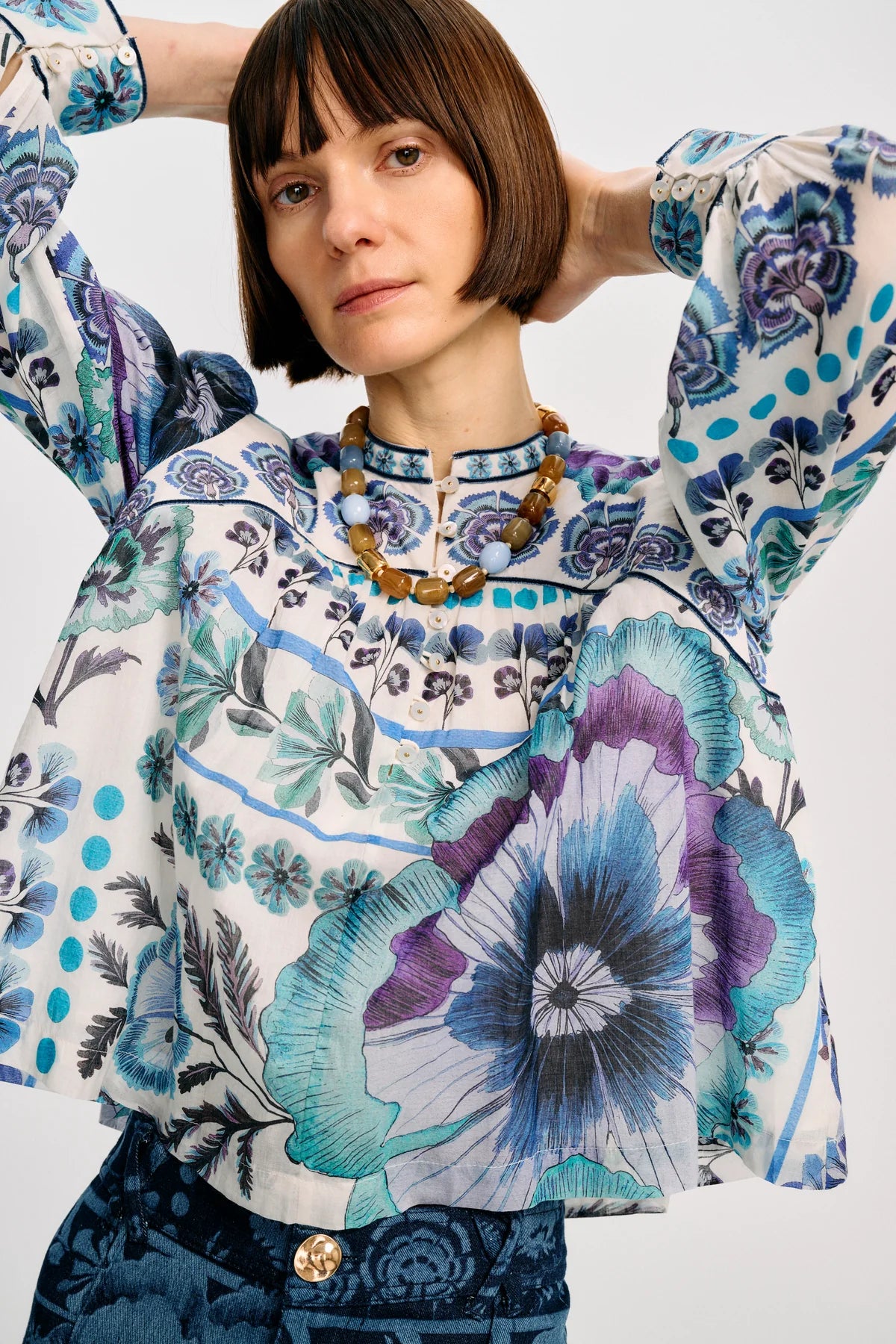 A model wearing a long-sleeve patterned blouse with blue, purple, and white floral motifs, paired with dark blue patterned trousers, against a light gray studio background.