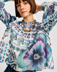 A model wearing a long-sleeve patterned blouse with blue, purple, and white floral motifs, paired with dark blue patterned trousers, against a light gray studio background.
