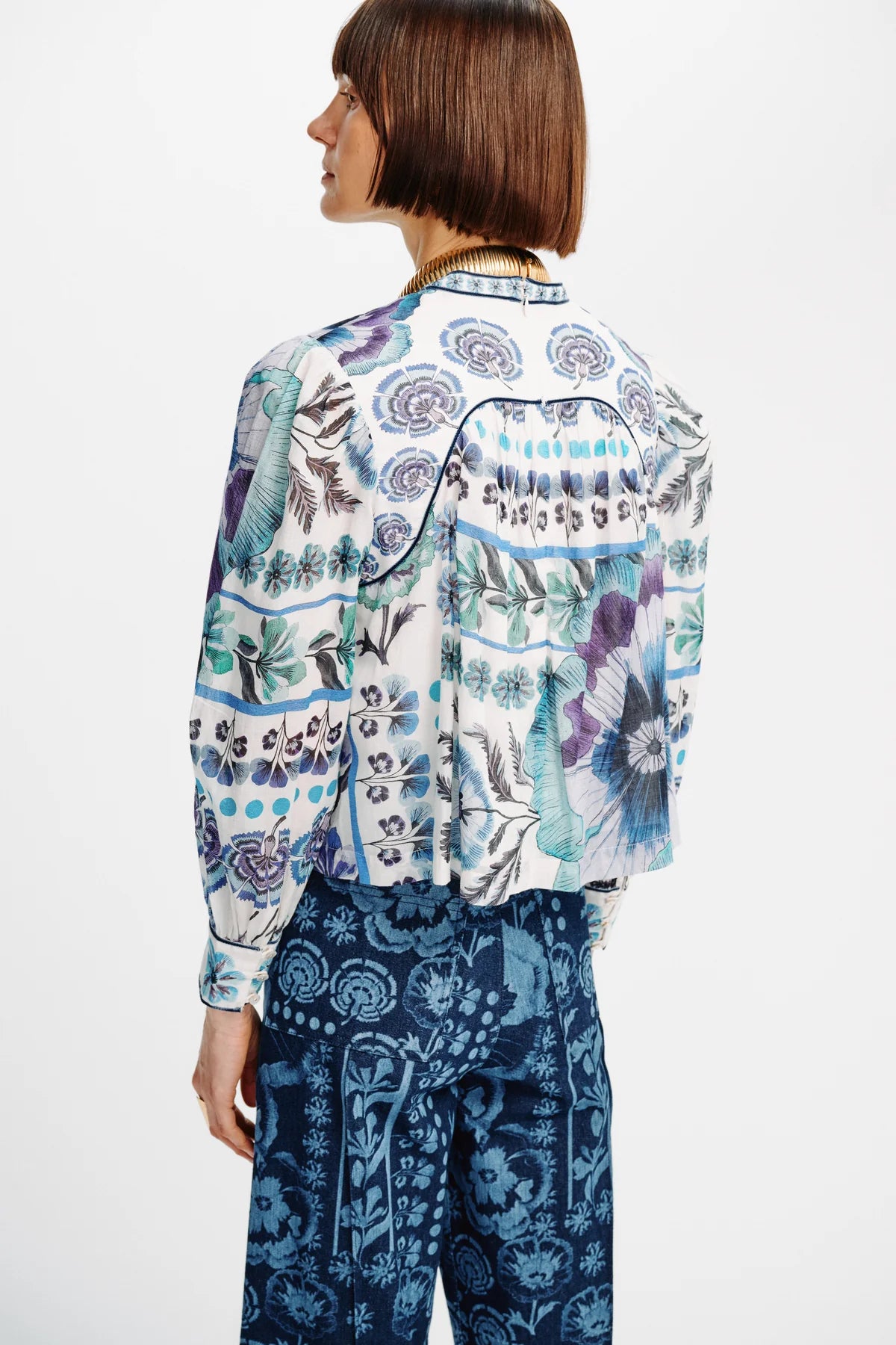 A model shown from the back wearing a white long-sleeve button-up shirt with a blue, purple, and green botanical pattern, paired with dark blue patterned trousers, against a plain white background.