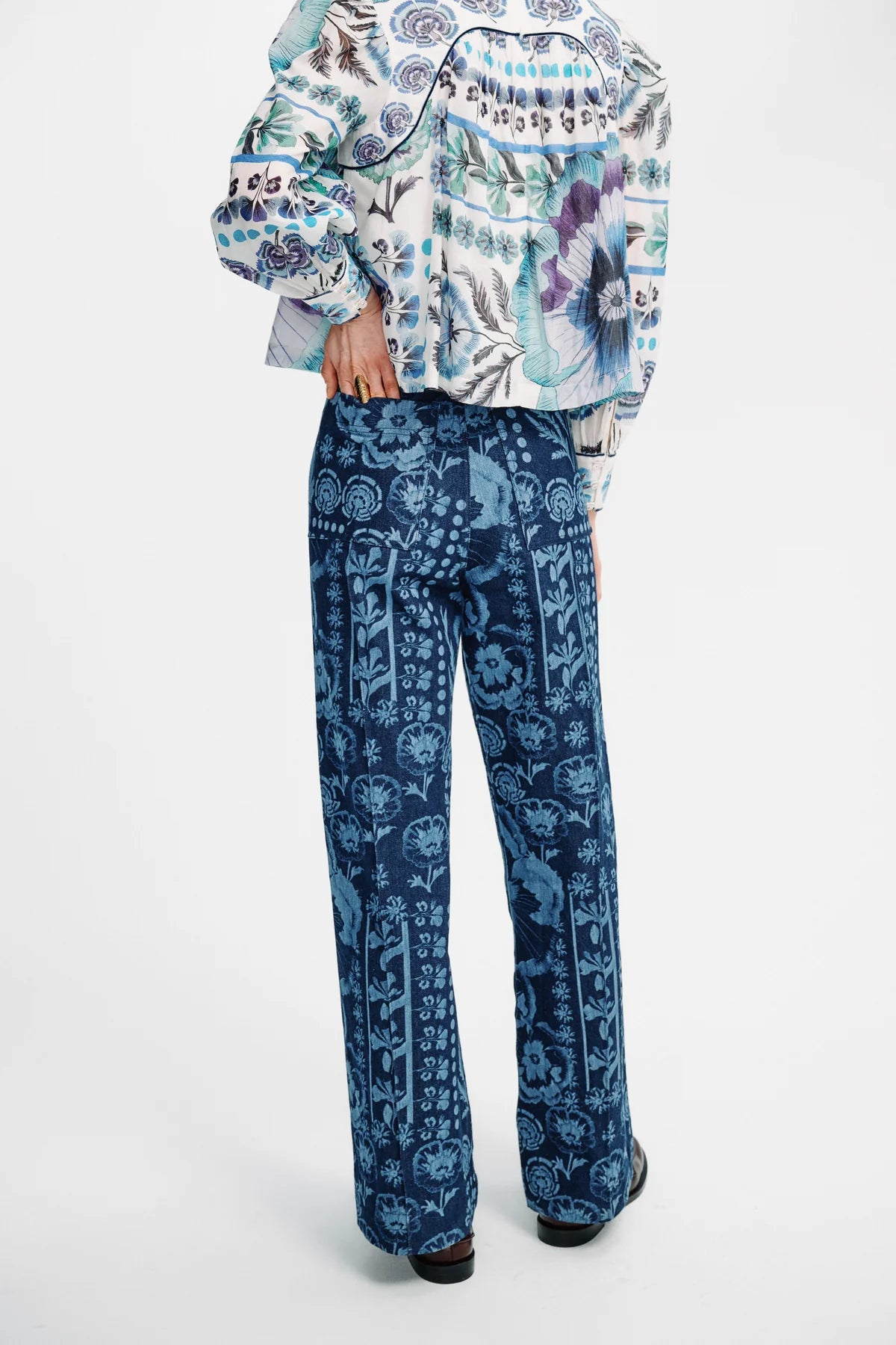 A model shown from the back wearing a patterned long-sleeve top and matching wide-leg trousers. The garments feature an intricate blue, white, and purple graphic print, photographed against a clean white studio background.