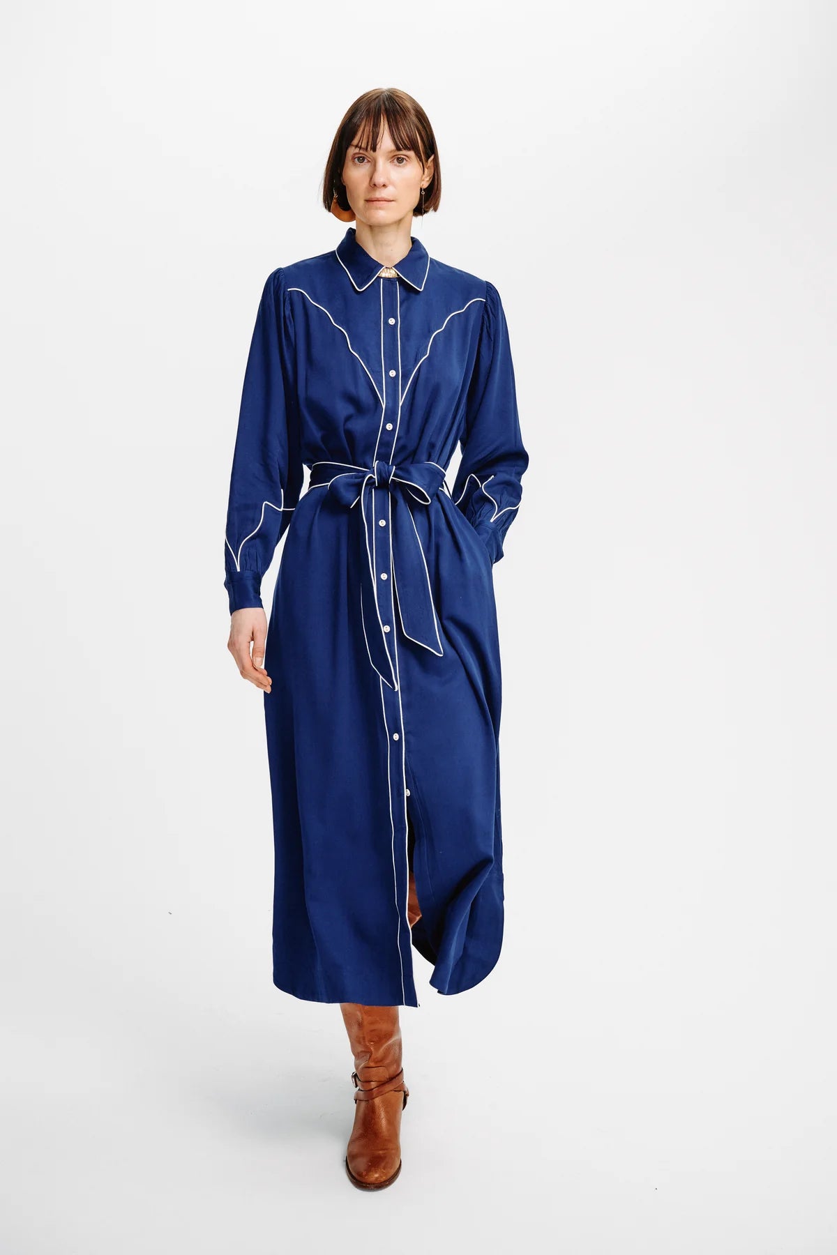 A model wearing a long-sleeved navy blue shirt dress with white contrast stitching and a matching tie belt, paired with brown leather boots, shot against a plain white studio background.