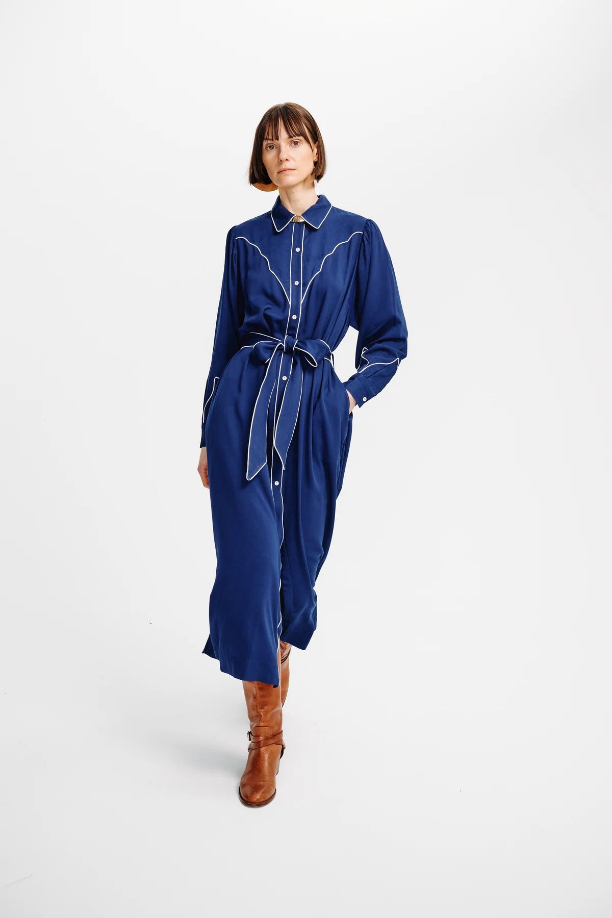 A model wearing a dark blue long-sleeve shirt dress with white contrast stitching and a matching tie belt, paired with brown leather boots, against a plain white studio background.