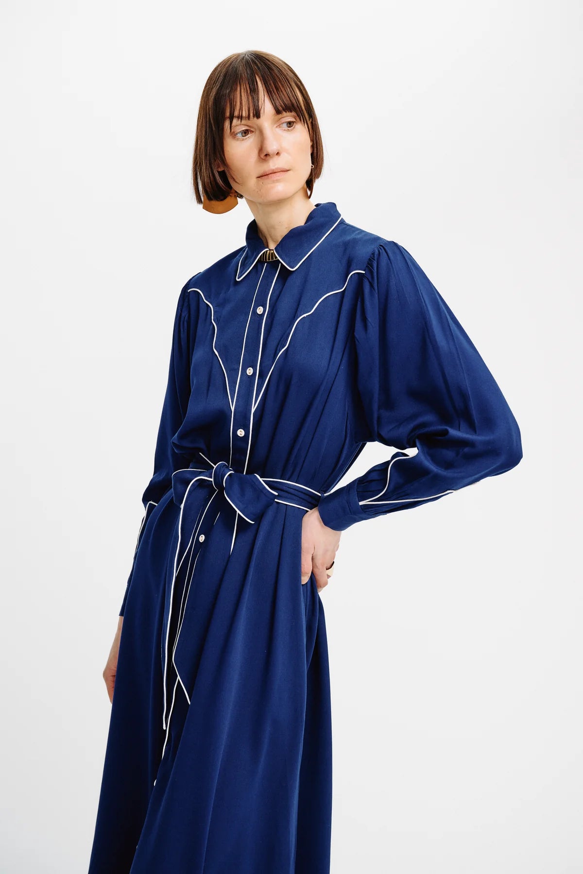 A woman wearing a long-sleeved navy blue shirt dress with white contrast stitching, a pointed collar, and a matching tie belt, photographed against a plain light gray studio background.