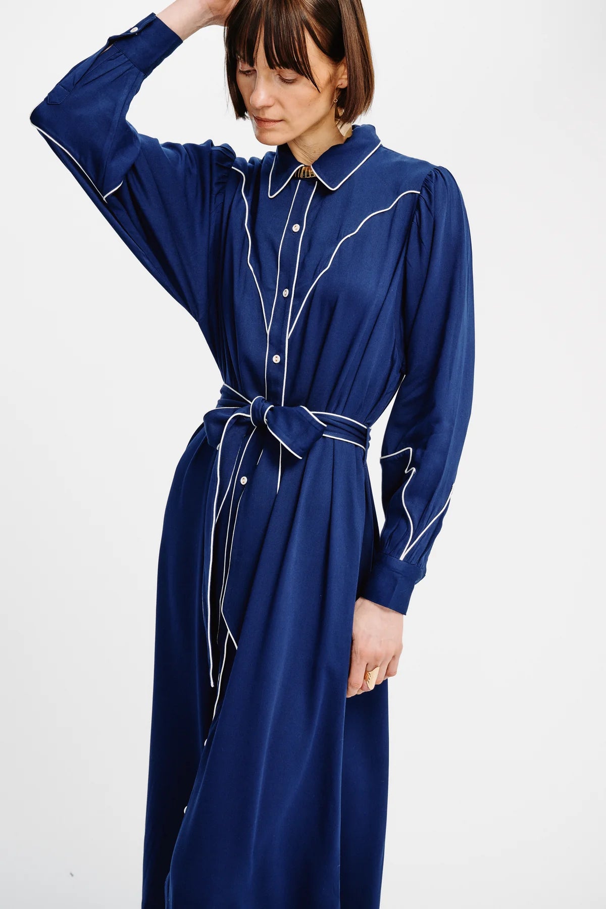 A woman wearing a long-sleeved navy blue shirt dress with white contrast piping and a matching tie belt, photographed against a plain white studio background.