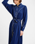 A woman wearing a long-sleeved navy blue shirt dress with white contrast piping and a matching tie belt, photographed against a plain white studio background.