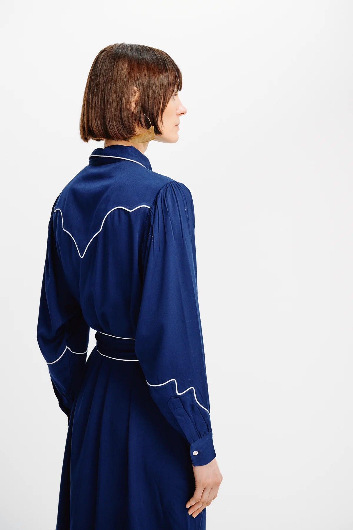 A woman with a short brown bob haircut is shown from the back wearing a navy blue long-sleeve dress with white contrast stitching details, set against a plain white studio background.