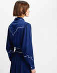 A woman with a short brown bob haircut is shown from the back wearing a navy blue long-sleeve dress with white contrast stitching details, set against a plain white studio background.