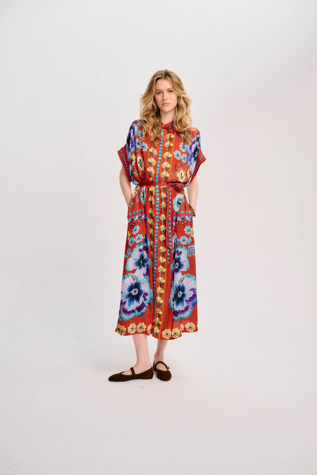 A woman modeling a long-sleeved, midi-length dress with a vibrant red base and a colorful floral and geometric print in blue, purple, yellow, and orange, paired with dark brown flat shoes, against a plain white studio background.