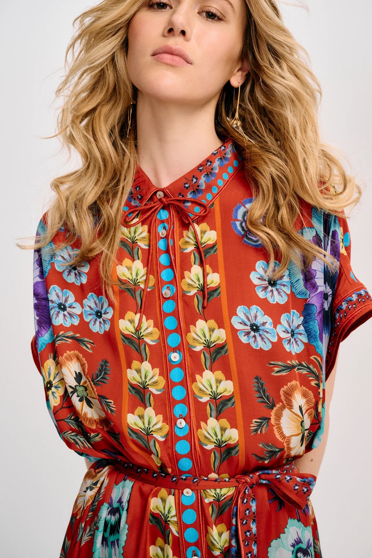 A close-up of a woman wearing a short-sleeve collared dress with a vibrant orange-red base and a multicolor floral print featuring yellow, blue, purple, and green flowers.