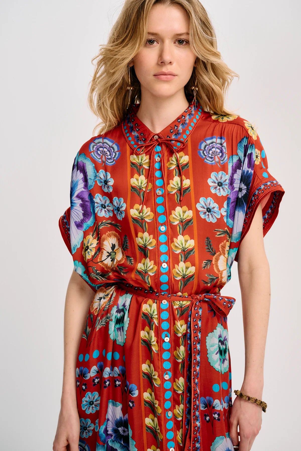 A woman modeling a short-sleeve, tie-neck dress with a vibrant red-orange base and a colorful floral print in blue, purple, yellow, and green, accented with turquoise buttons down the front.