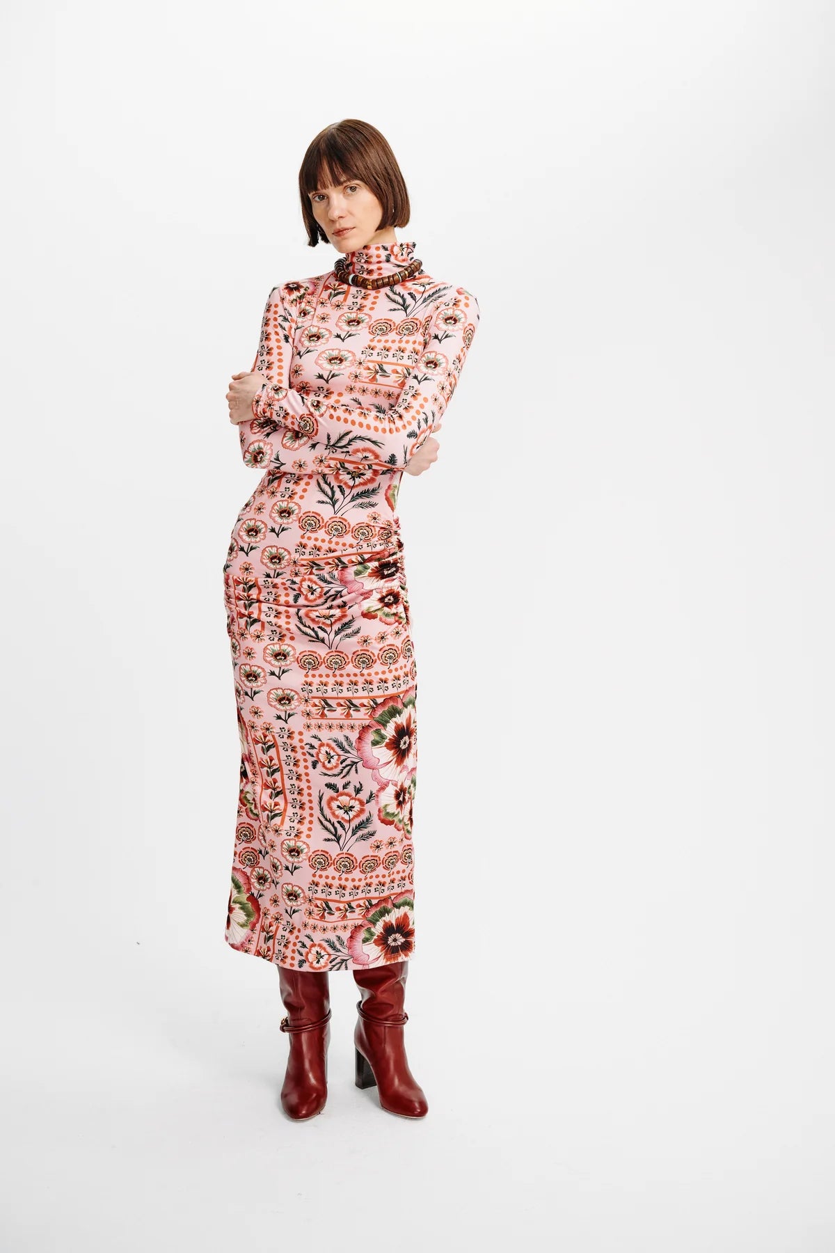 A model wearing a long-sleeved mock-neck midi dress with a colorful geometric and tribal-inspired pattern in pink, orange, red, and black, paired with dark red ankle boots, against a plain white studio background.