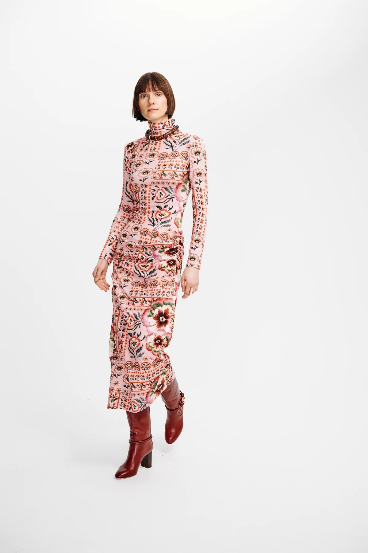 A model wearing a long-sleeved, high-neck midi dress with a pink and beige multicolor paisley and floral pattern, paired with dark red ankle boots, against a plain white studio background.