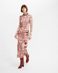 A model wearing a long-sleeved, high-neck midi dress with a pink and beige multicolor paisley and floral pattern, paired with dark red ankle boots, against a plain white studio background.