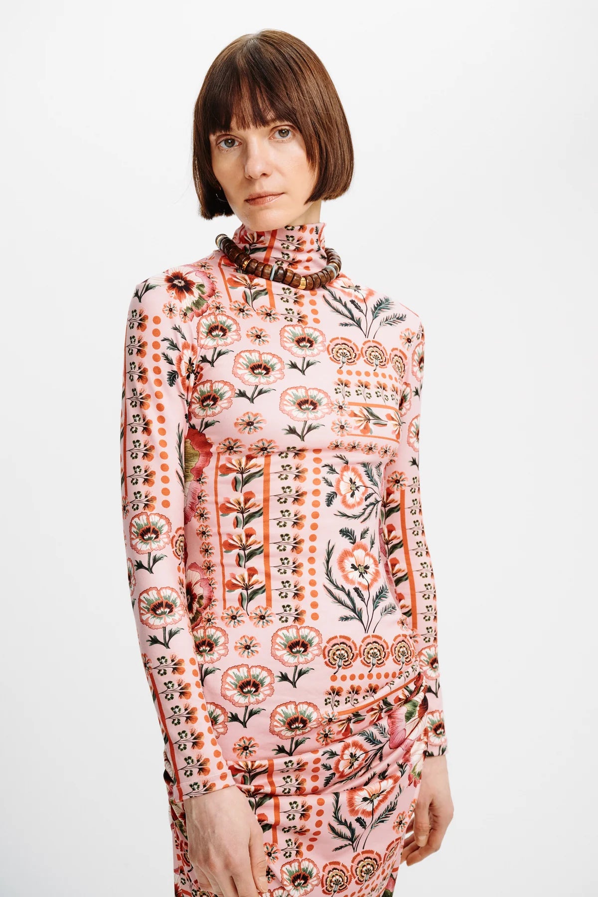 A woman with a short brown bob haircut wearing a long-sleeve mock-neck dress with a pink base and a detailed multicolor floral and geometric pattern, photographed against a plain white studio background.