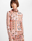 A woman with a short brown bob haircut wearing a long-sleeve mock-neck dress with a pink base and a detailed multicolor floral and geometric pattern, photographed against a plain white studio background.