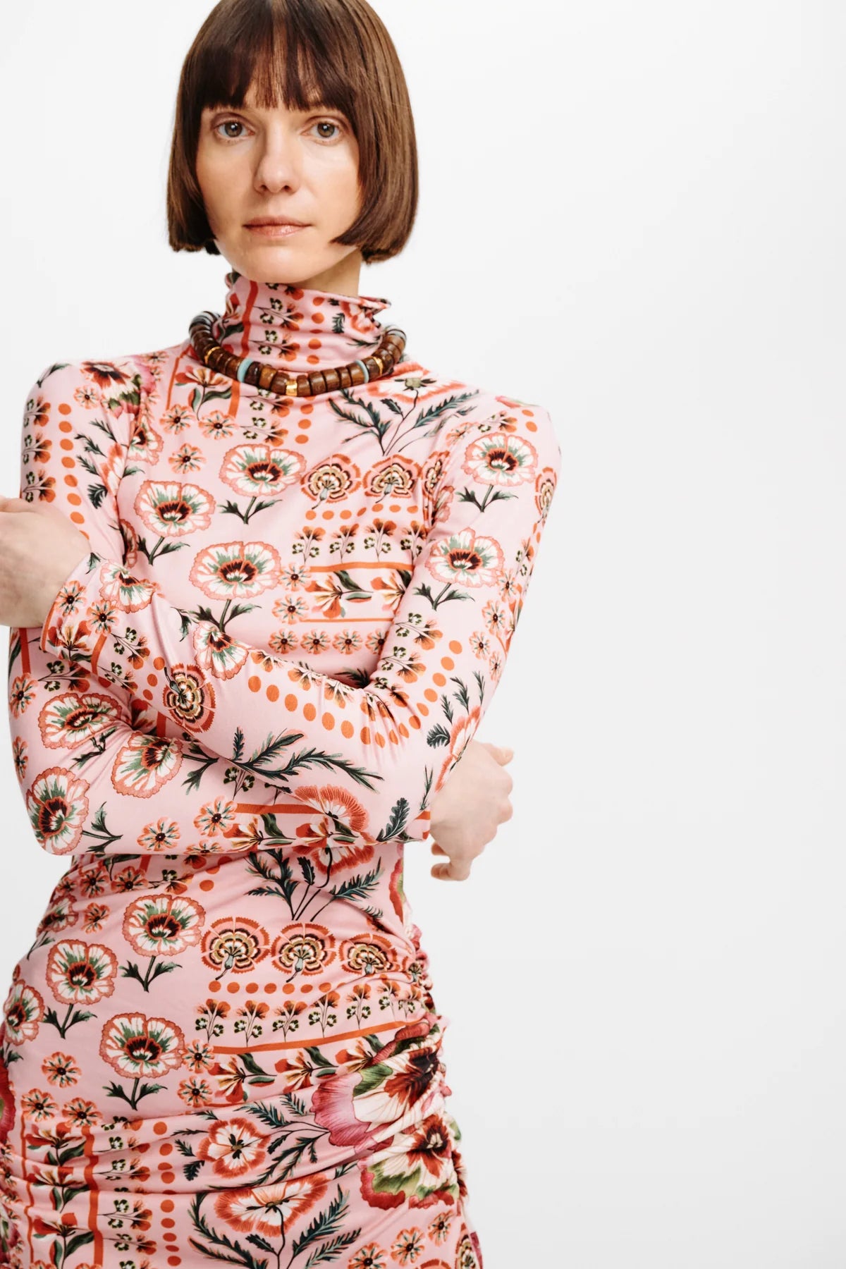 A model with a short brown bob haircut wearing a pink mock-neck dress with a dense orange, red, and black floral and botanical pattern, posed against a solid light gray background.