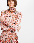 A model with a short brown bob haircut wearing a pink mock-neck dress with a dense orange, red, and black floral and botanical pattern, posed against a solid light gray background.
