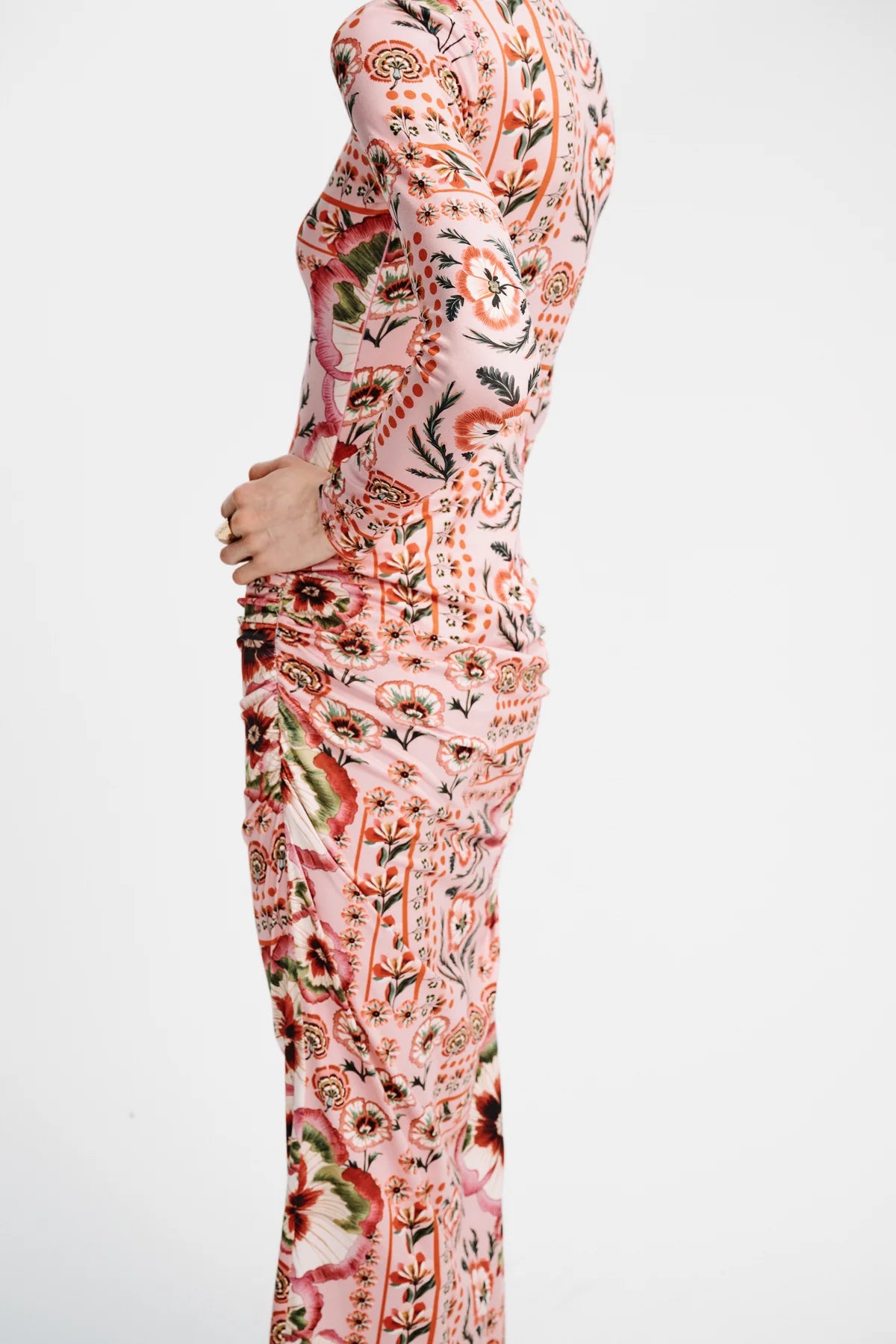 A model shown from the side wearing a long-sleeve, high-neck maxi dress with a dense botanical and paisley print in pink, orange, green, and black on a white background.