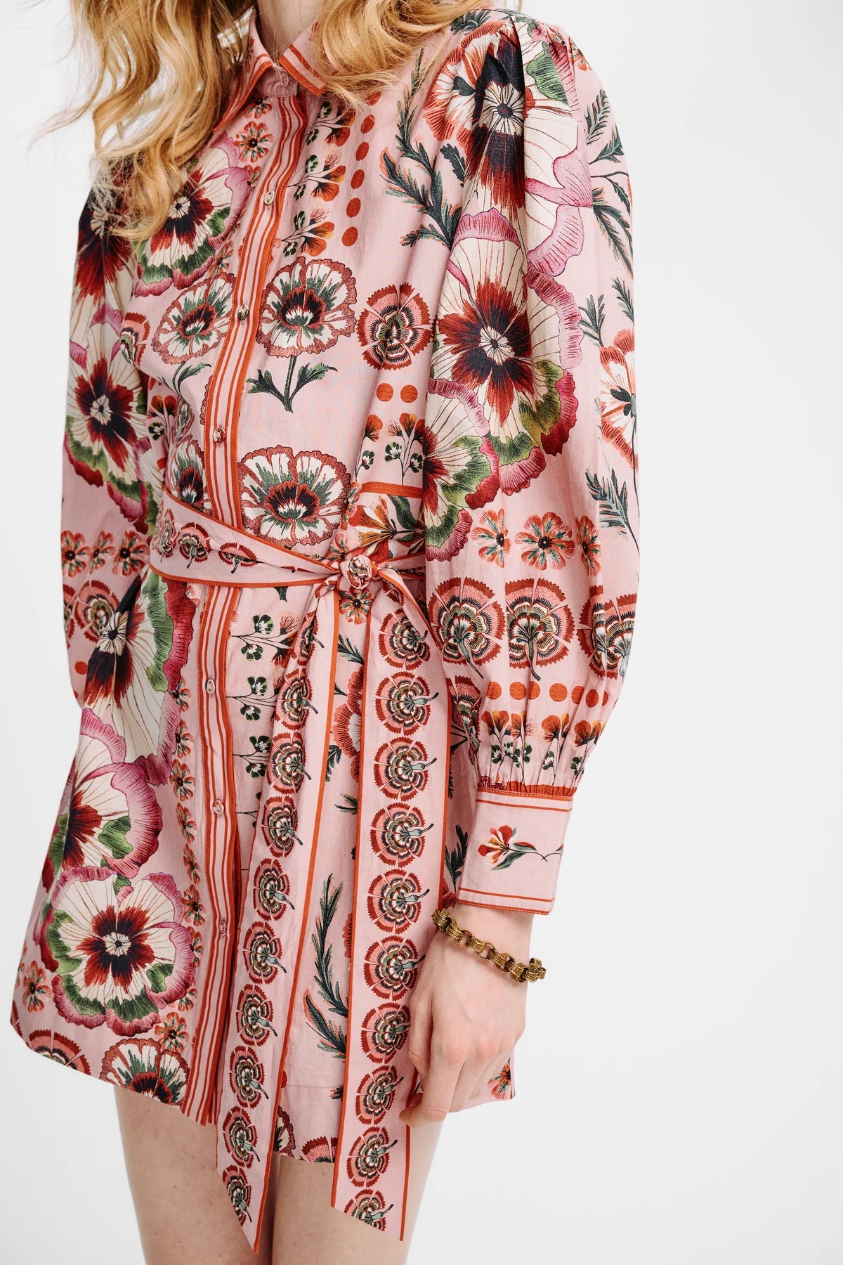 A close-up of a model wearing a pink long-sleeve shirt dress with a bold floral and geometric print in red, green, and black, featuring a tie waist belt.
