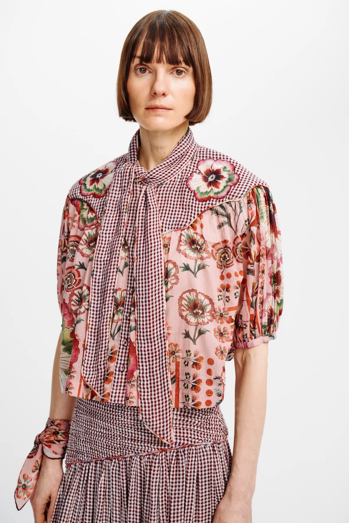 A model wearing a multi-patterned blouse with pink floral prints, red-and-white gingham, and puff sleeves, paired with a matching gingham skirt against a plain white background.