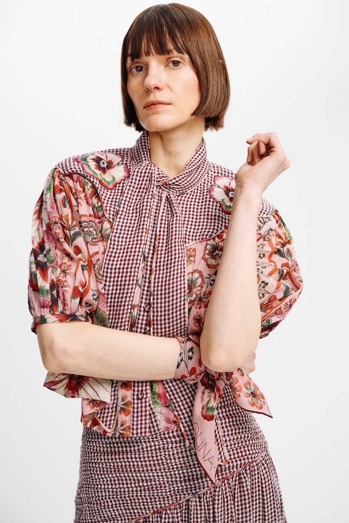 A woman with a short brown bob haircut wearing a pink and white gingham blouse with floral puff sleeves and a tie-neck detail, paired with a matching gingham skirt, against a plain light gray background.