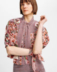 A woman with a short brown bob haircut wearing a pink and white gingham blouse with floral puff sleeves and a tie-neck detail, paired with a matching gingham skirt, against a plain light gray background.