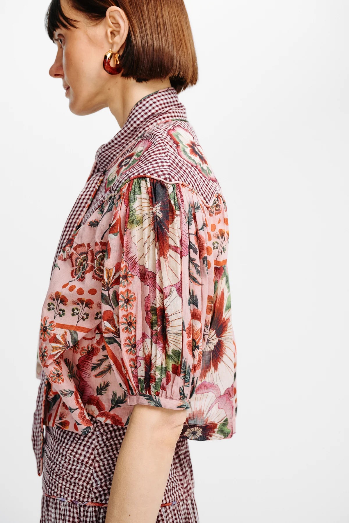 A side-profile view of a model wearing a short-sleeved blouse with a pink floral print and a red-and-white gingham collar, paired with a matching gingham skirt against a plain white background.