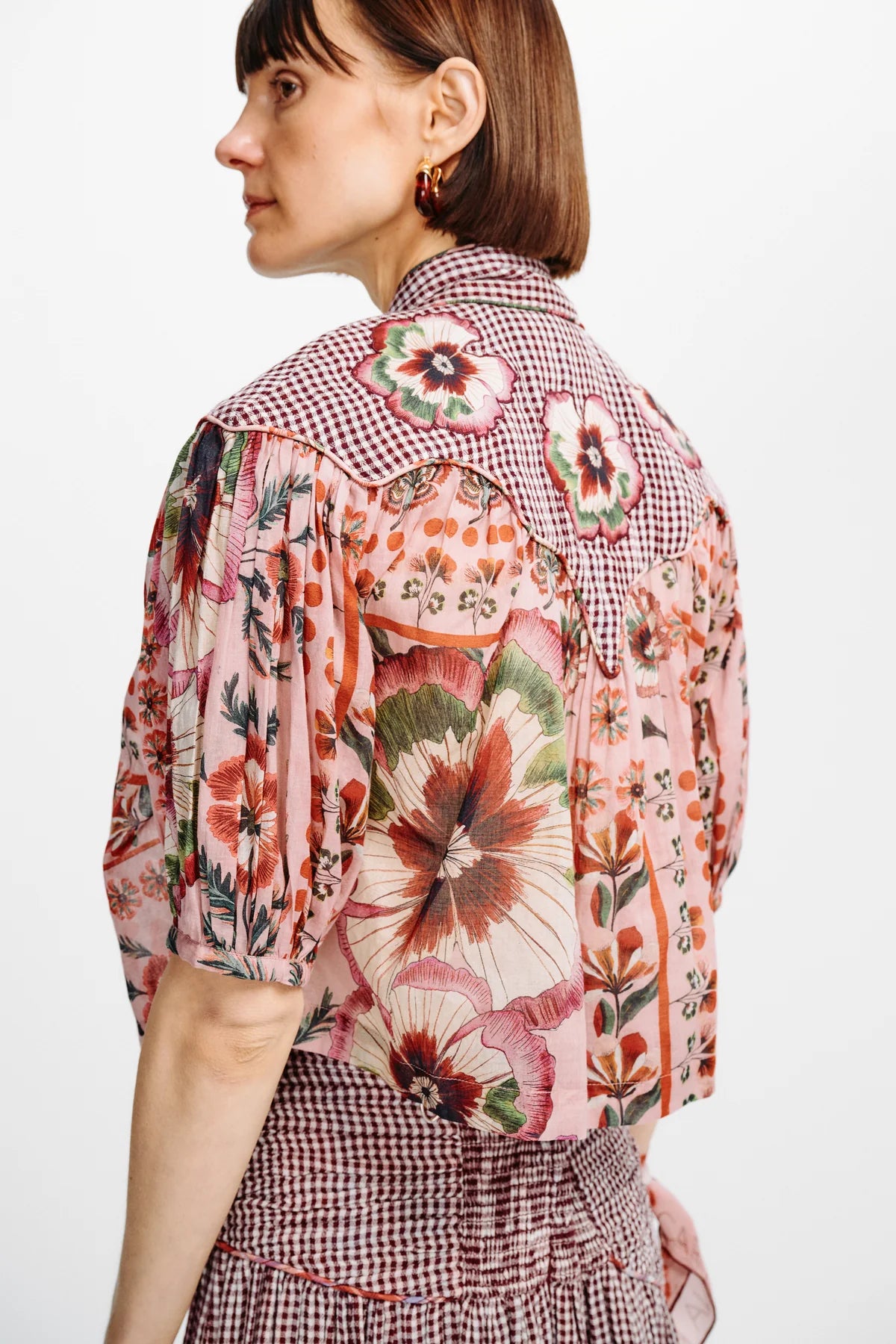 A woman shown from the back wearing a short-sleeve button-front blouse with a mixed floral and gingham print in pink, red, green, and beige tones, paired with a matching gingham skirt against a plain light background.