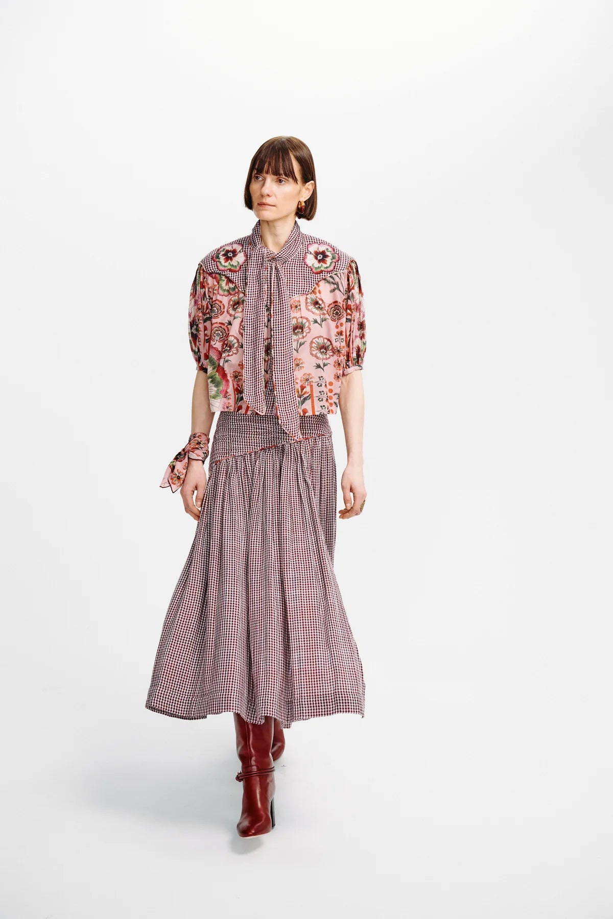 A model walks against a light gray studio background wearing a coordinated outfit consisting of a dusty rose and pink plaid midi skirt, a matching tie-neck blouse with heavily embroidered floral sleeves, and dark red pointed boots.