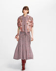 A model walks against a light gray studio background wearing a coordinated outfit consisting of a dusty rose and pink plaid midi skirt, a matching tie-neck blouse with heavily embroidered floral sleeves, and dark red pointed boots.