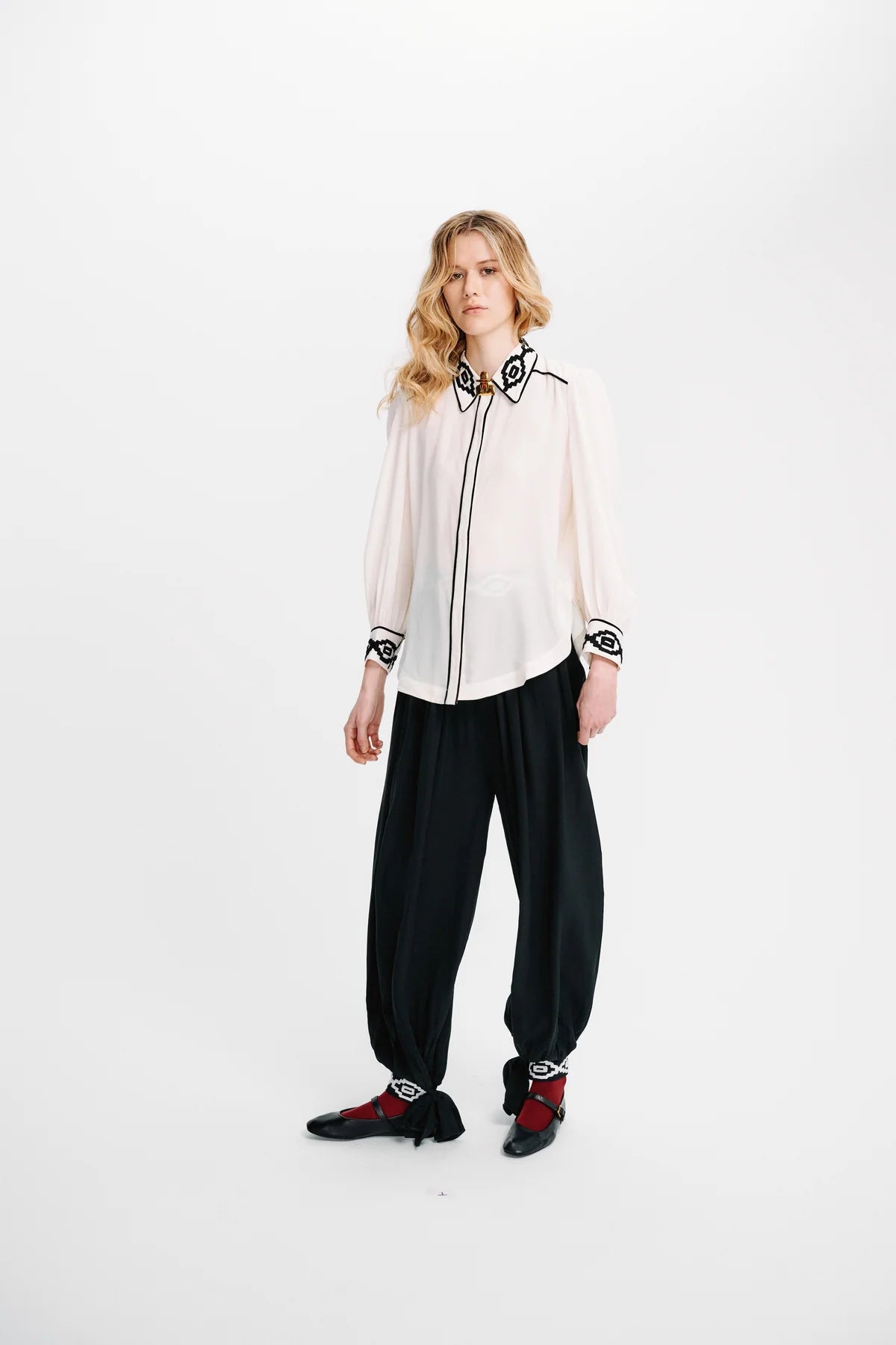 A model stands against a plain white studio background wearing a white long-sleeve blouse with black embroidered trim, black wide-leg pants, and black shoes with red accents.