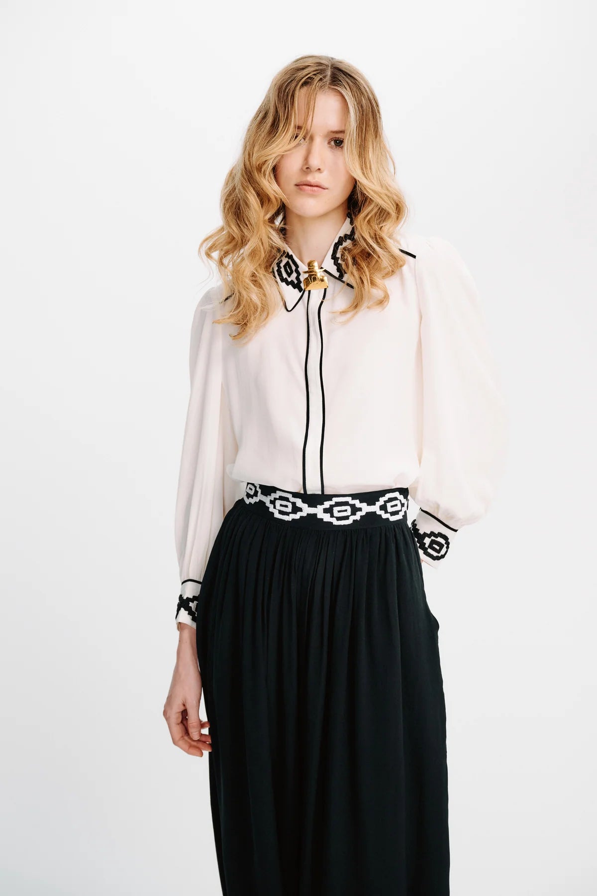 A woman modeling a sheer off-white blouse with black embroidered trim and a black skirt with a black-and-white patterned belt, against a plain white studio background.