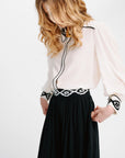 A woman wearing a cream-colored blouse with black trim and a black skirt, shown from the side against a plain white background.