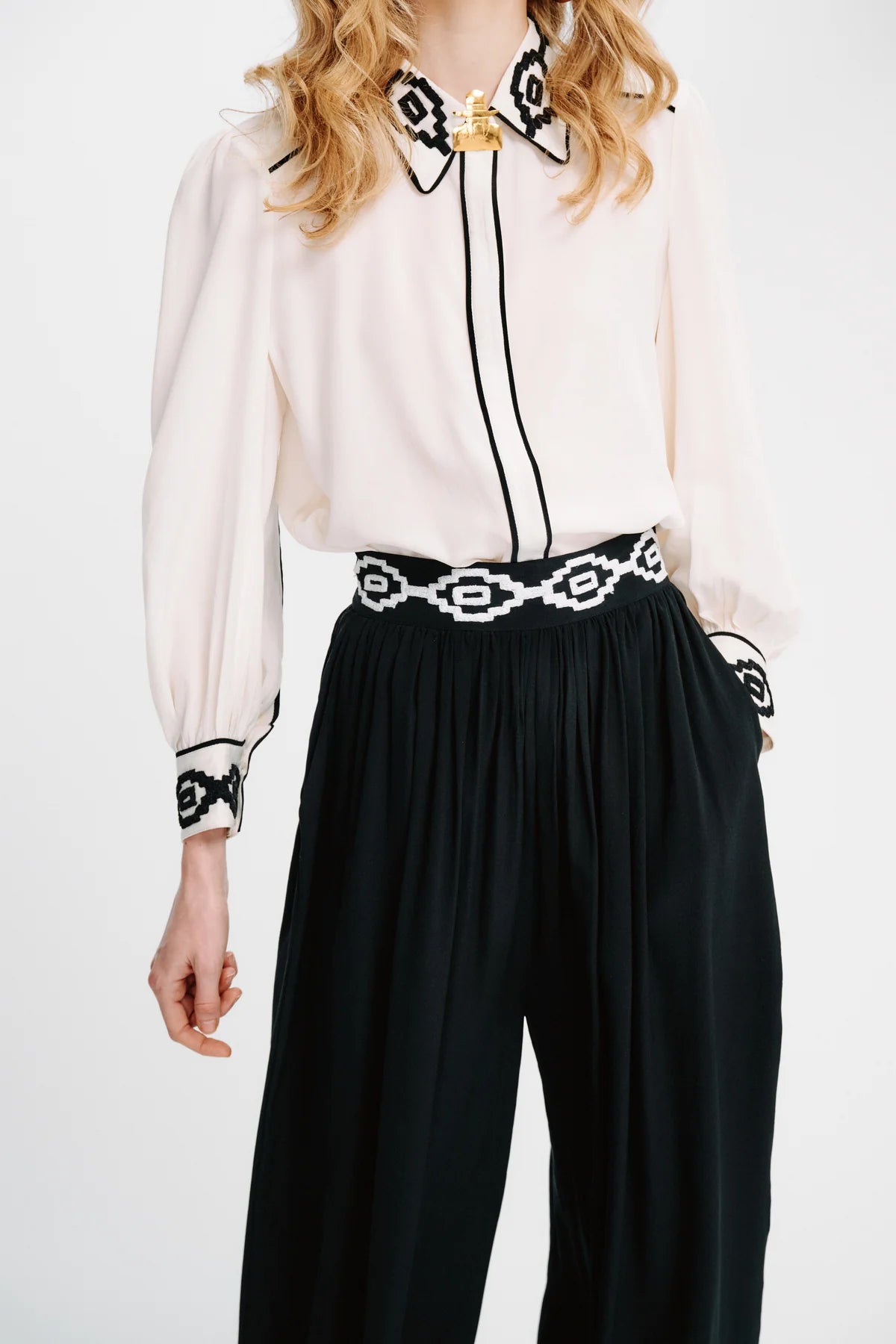 A woman wearing a cream long-sleeve blouse with black and white patterned collar, cuffs, and center placket, paired with black wide-leg pants featuring a matching black and white chain-link waistband.