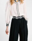 A woman wearing a cream long-sleeve blouse with black and white patterned collar, cuffs, and center placket, paired with black wide-leg pants featuring a matching black and white chain-link waistband.