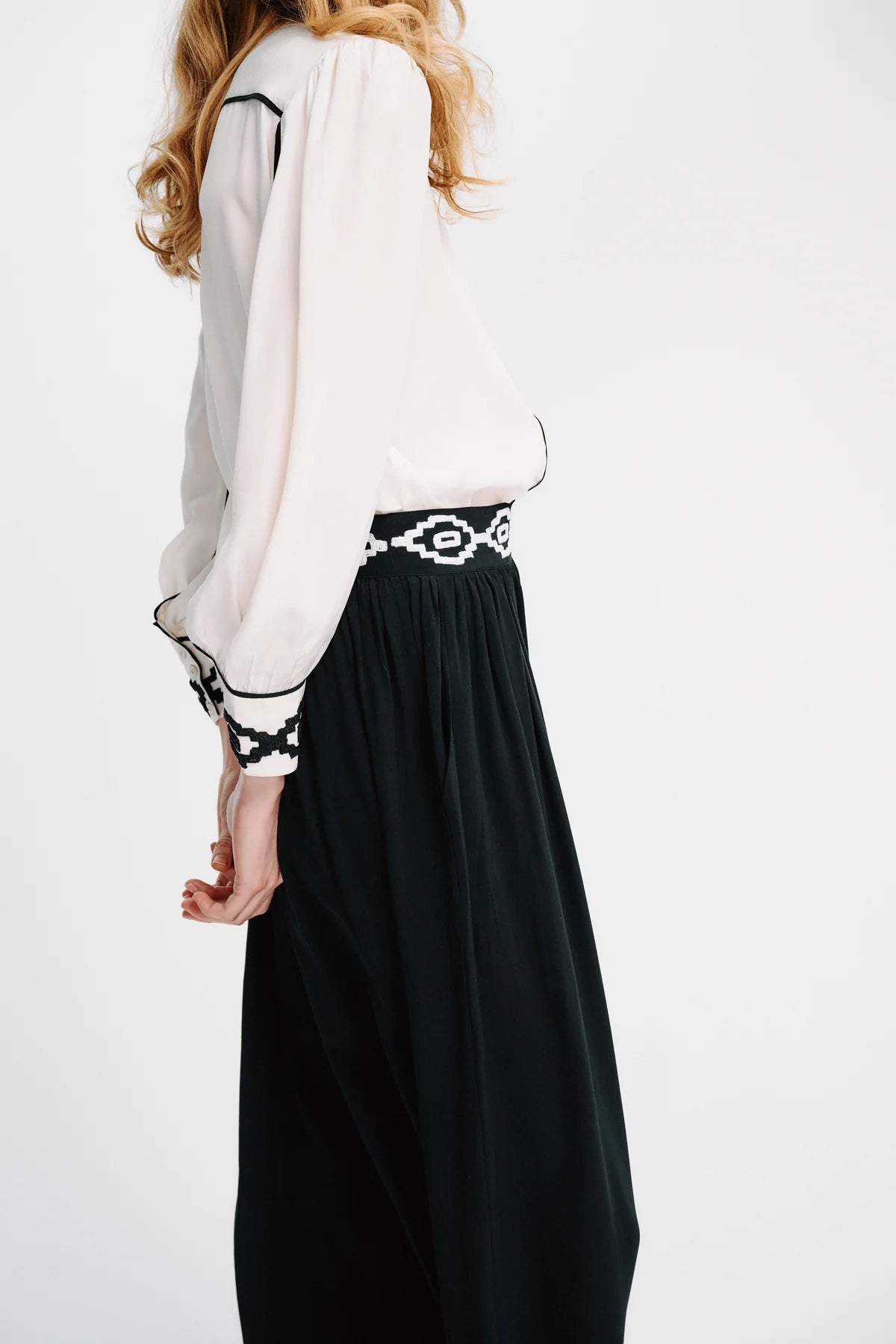 A woman shown from the back wearing a white blouse with black embroidered trim on the cuffs and a black skirt with a black and white patterned waistband, against a plain white studio background.