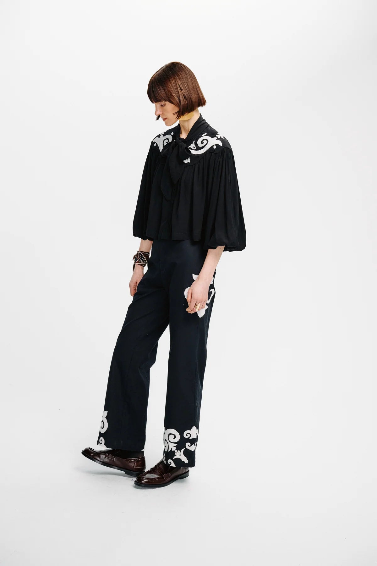 A model wearing a black long-sleeve blouse with white baroque-style embroidery on the shoulders and a matching black wide-leg trouser set with white embroidery on the cuffs, paired with dark brown leather loafers, against a plain white studio background.