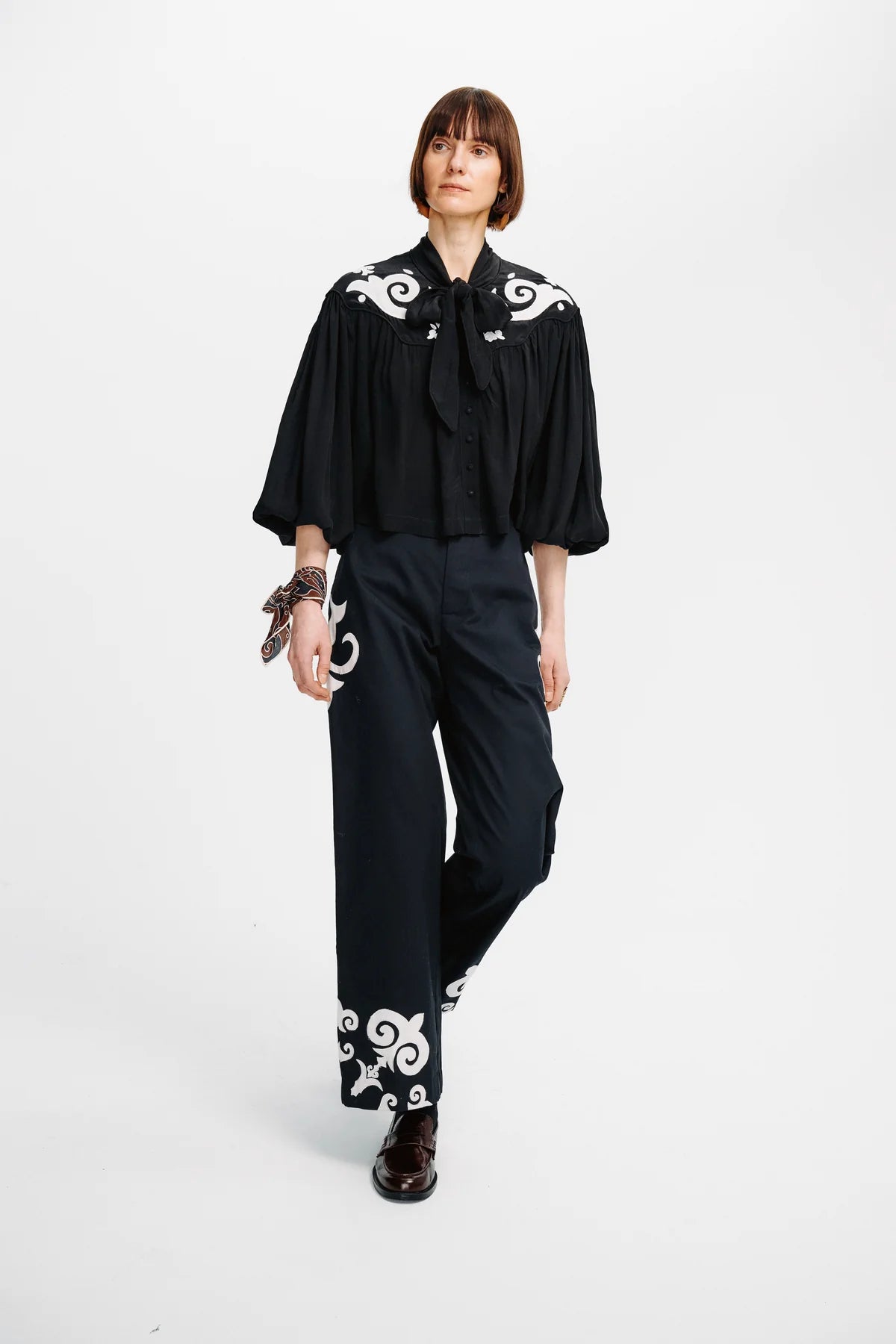 A model wearing a black blouse with white scroll embroidery on the shoulders and sleeves, paired with matching black wide-leg trousers featuring white embroidery near the hem, set against a plain white studio background.