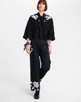 A model wearing a black blouse with white scroll embroidery on the shoulders and sleeves, paired with matching black wide-leg trousers featuring white embroidery near the hem, set against a plain white studio background.