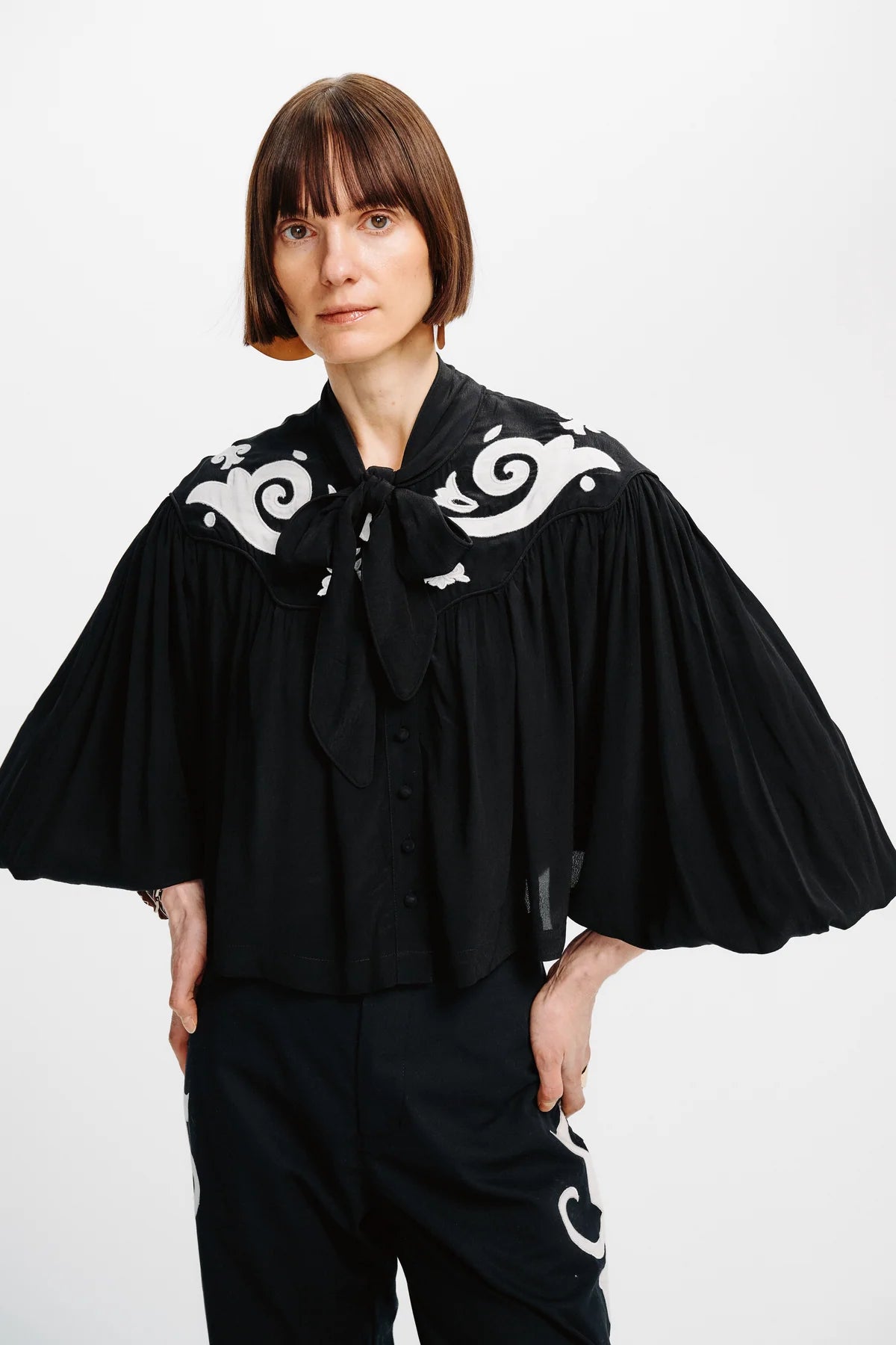 A model wearing a black blouse with dramatic pleated bell sleeves, a large bow tie-neck, and white scrollwork embroidery across the shoulders, paired with black pants against a plain white studio background.