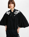 A model wearing a black blouse with dramatic pleated bell sleeves, a large bow tie-neck, and white scrollwork embroidery across the shoulders, paired with black pants against a plain white studio background.