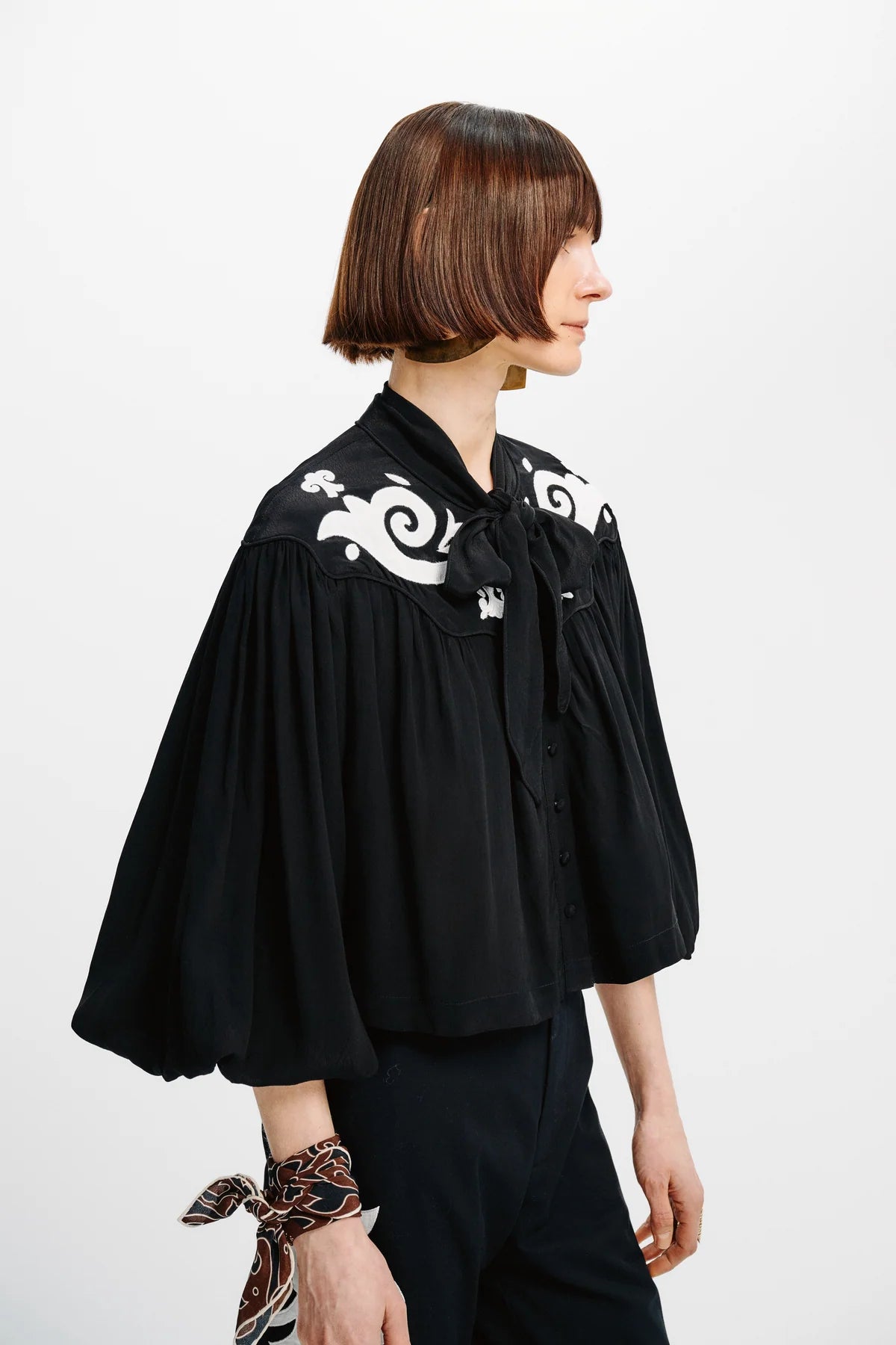 A woman shown in profile wearing a black cropped blouse with dramatic puffed sleeves and white baroque-style embroidery on the shoulders, paired with dark trousers against a plain light background.