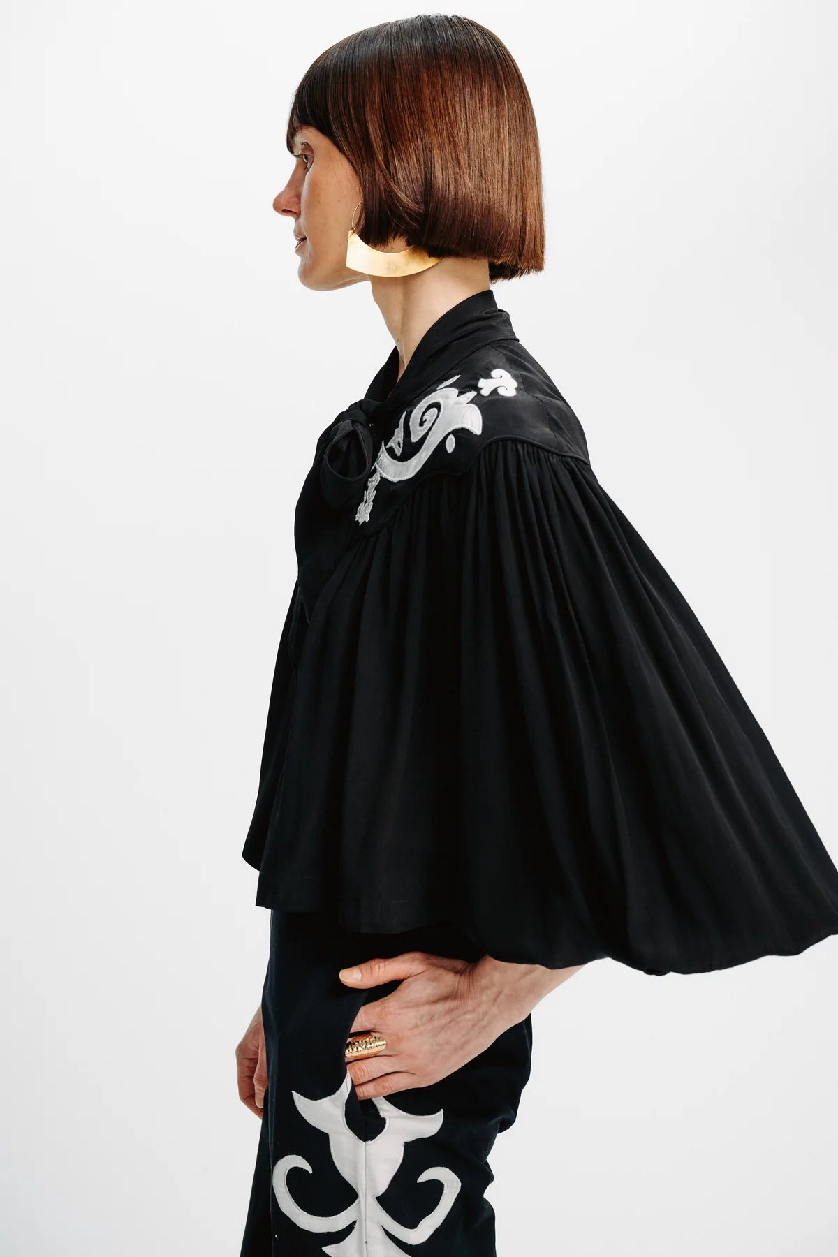 A model in profile wearing a black pleated blouse with white embroidered details on the shoulder and a matching black skirt with white embroidery, set against a plain white studio background.